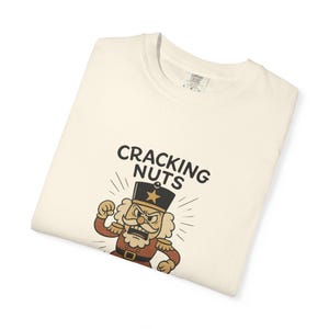 Vintage style Christmas shirt, with rubber hose style nut cracker cartoon character, with "Cracking Nuts and Taking Names" caption, on folded ivory comfort colors tee.