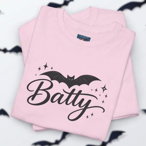 Puede incluir: Una camiseta de color rosa claro con un gr&aacute;fico negro de un murci&eacute;lago sobre la palabra Batty en letra cursiva. El dise&ntilde;o incluye peque&ntilde;os detalles de estrellas negras alrededor del murci&eacute;lago y el texto, con una etiqueta azul en el cuello.
