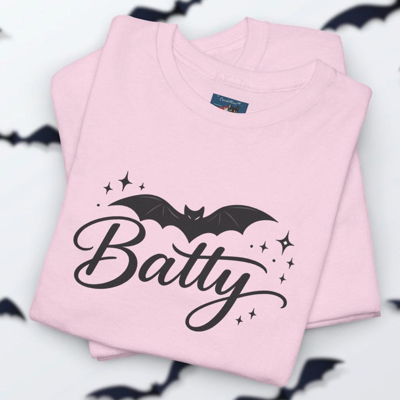 Puede incluir: Una camiseta de color rosa claro con un gr&aacute;fico negro de un murci&eacute;lago sobre la palabra Batty en letra cursiva. El dise&ntilde;o incluye peque&ntilde;os detalles de estrellas negras alrededor del murci&eacute;lago y el texto, con una etiqueta azul en el cuello.