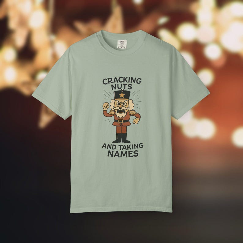 Vintage style Christmas shirt, with rubber hose style nut cracker cartoon character, with "Cracking Nuts and Taking Names" caption, on pastel green comfort colors tee.