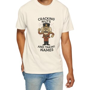 Vintage style Christmas shirt, with rubber hose style nut cracker cartoon character, with "Cracking Nuts and Taking Names" caption, on model wearing ivory comfort colors tee.