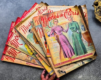Op de afbeelding: Een stapel vintage "Modes de Paris" tijdschriften. De covers tonen illustraties van vrouwen in jurken, met Franse tekst. De tijdschriften hebben een versleten, verouderd uiterlijk, met rode en gele tinten.