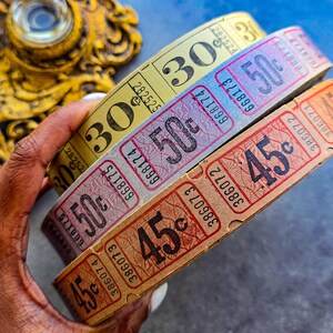 May include: Three vintage ticket rolls in yellow, blue, and orange. Each roll displays a large number: 30, 50, and 45, respectively. The tickets have a retro aesthetic with serial numbers and a faded appearance, suggesting age and use.