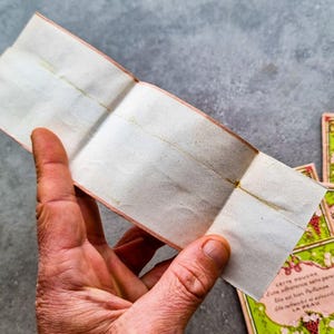 May include: A folded piece of paper with a pink border and a faint yellow line down the center. The paper is blank and appears to be old. The text on the bottom of the image reads "CETTE POUDRE d'une adherence sans pares Gile est bien Parfla Alle rafraichit at adoucit LA PEAU".
