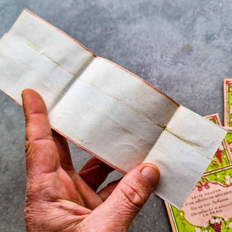 May include: A folded piece of paper with a pink border and a faint yellow line down the center. The paper is blank and appears to be old. The text on the bottom of the image reads "CETTE POUDRE d'une adherence sans pares Gile est bien Parfla Alle rafraichit at adoucit LA PEAU".
