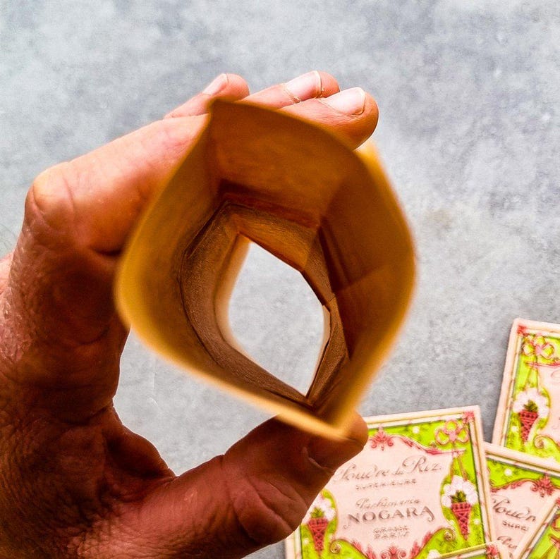 May include: A brown paper bag is folded into a cone shape, revealing a glimpse of a colorful patterned box with the text "Cr&egrave;me de Riz" and "Nogara" visible. The box features a pink and green design with a floral pattern and a small image of a strawberry.