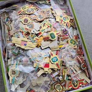 May include: A collection of assorted paper cigar bands in a green box, each featuring unique colourful illustrations, gold foil accents, and various printed text labels, individually housed in small clear plastic bags for preservation and organisation.