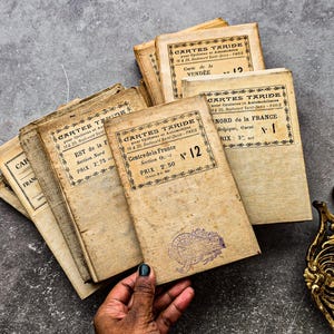 Op de afbeelding: Een verzameling vintage Franse reisgidsen, met verouderd papier en vervaagde inkt. De covers tonen de titel "CARTES TARIDE" met regionale namen en nummers. De boeken zijn gestapeld op een grijze, getextureerde ondergrond.