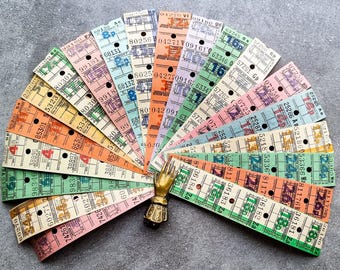 Op de afbeelding: Een verzameling vintage papieren tickets in verschillende kleuren, gerangschikt in een waaier. De tickets hebben gedrukte nummers en letters, en sommige hebben kleine gaatjes die er doorheen zijn geponst. Een goudkleurig handvormig decoratief object is in het midden van de waaier geplaatst.