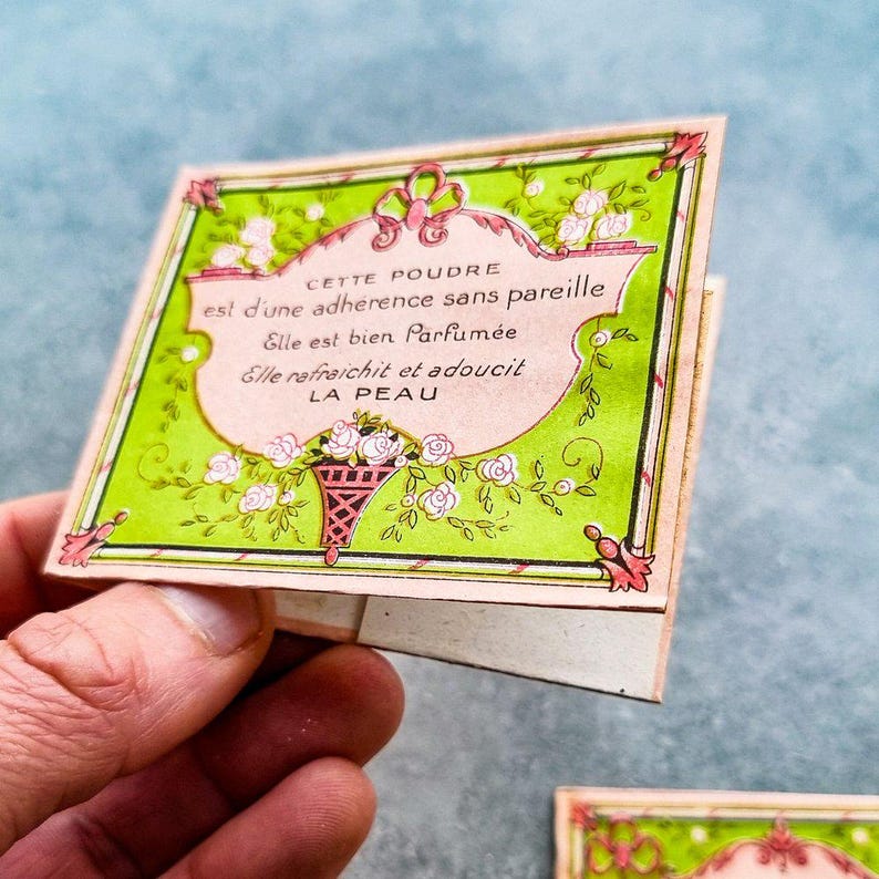 May include: A vintage pink and green powder compact with a floral design and the text "CETTE POUDRE est d'une adherence sans pareille Elle est bien Parfum&eacute;e Elle rafraichit et adoucit LA PEAU".