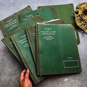 Op de afbeelding: Een verzameling vintage groene boeken met de tekst "SOCIETE DE FONCAGE DE PUITS FRANCO-BELGE BRUXELLES" op de kaft. De boeken zijn op een grijze ondergrond gestapeld, met een decoratief gouden object op de achtergrond.