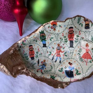 Pode incluir: Um prato decorativo de formato irregular com uma borda pintada de ouro. O interior apresenta um fundo verde claro com um padrão repetido de quebra-nozes, ratos e bailarinas. Enfeites de Natal no fundo.