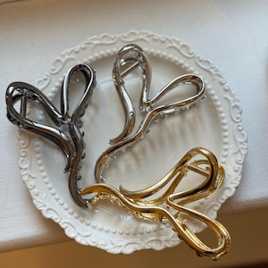 May include: Three metal hair claw clips in silver, gold, and gunmetal grey, arranged on a white, decorative plate. The clips have a curved, open design with a spring mechanism. The gold clip has a shiny finish, while the silver clip has small, clear embellishments.