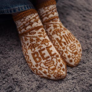 Peut inclure: Une paire de chaussettes tricotées présentant un motif blanc et brun doré. Le design inclut des motifs floraux, le texte "BEE" sur une chaussette et "KIND" sur l'autre, accompagnés de petites illustrations d'abeilles et de bords-côtes.