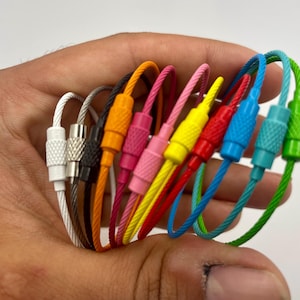 May include: A collection of circular wire cable keychains featuring screw-lock barrel connectors in various colors including white, silver, black, orange, pink, yellow, red, blue, teal, and green, arranged in a fan-like formation.