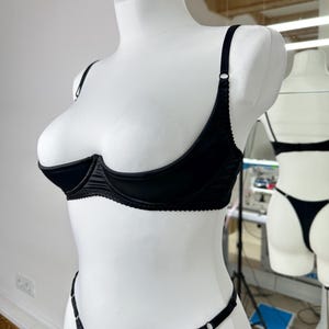 Peut inclure: Ensemble de lingerie noir pr&eacute;sent&eacute; sur un mannequin blanc. L'ensemble comprend un soutien-gorge demi-bonnet avec des bretelles r&eacute;glables et un string assorti. Le soutien-gorge et le string sont orn&eacute;s de d&eacute;tails d&eacute;licats. La lingerie est en tissu lisse et brillant.