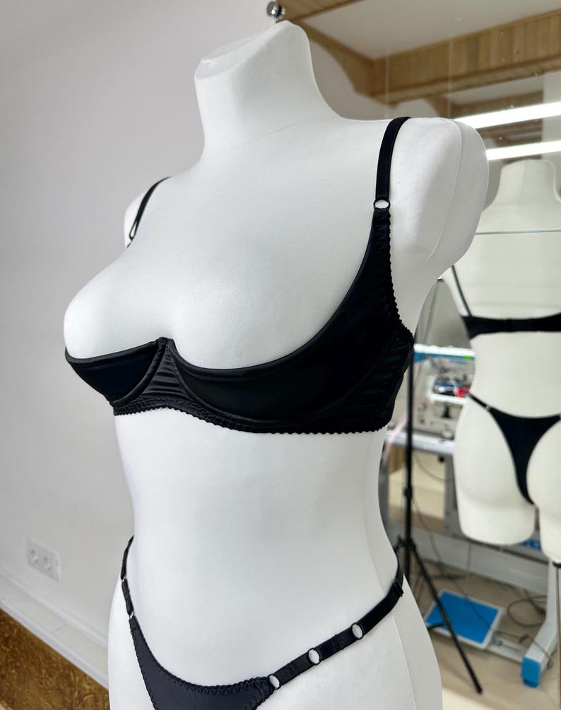 Peut inclure: Ensemble de lingerie noir pr&eacute;sent&eacute; sur un mannequin blanc. L'ensemble comprend un soutien-gorge demi-bonnet avec des bretelles r&eacute;glables et un string assorti. Le soutien-gorge et le string sont orn&eacute;s de d&eacute;tails d&eacute;licats. La lingerie est en tissu lisse et brillant.