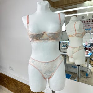 May include: A two-piece lingerie set featuring a lace underwire bra and matching high-cut panties. The set is crafted from cream-colored lace with a subtle blue floral pattern and is accented with thin, solid peach-colored straps and trim.