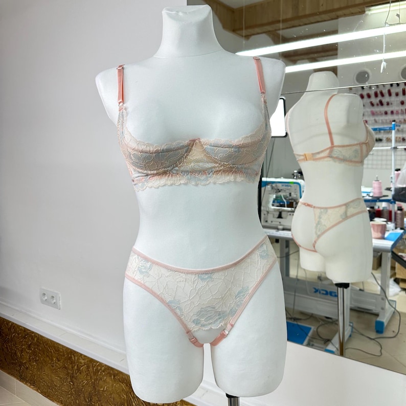 May include: A two-piece lingerie set featuring a lace underwire bra and matching high-cut panties. The set is crafted from cream-colored lace with a subtle blue floral pattern and is accented with thin, solid peach-colored straps and trim.