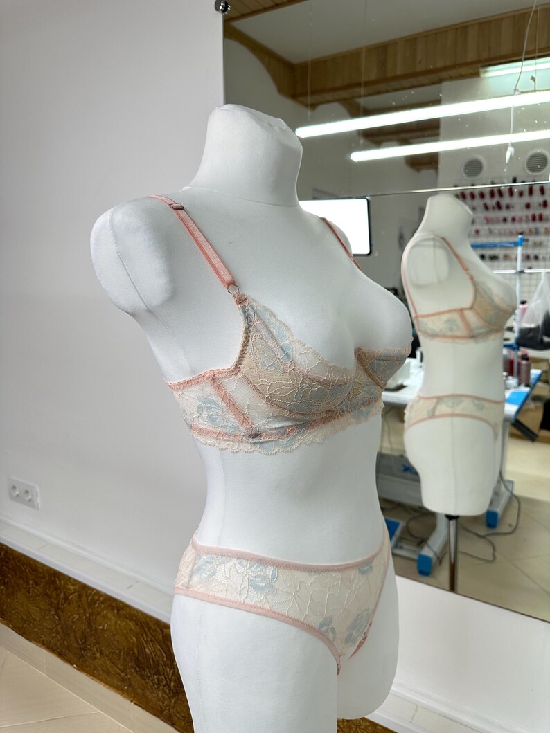 May include: A beige and light blue lace lingerie set, including a bra and matching panties, displayed on a white mannequin. The bra features adjustable pink straps and delicate lace detailing. The set is reflected in a nearby mirror.