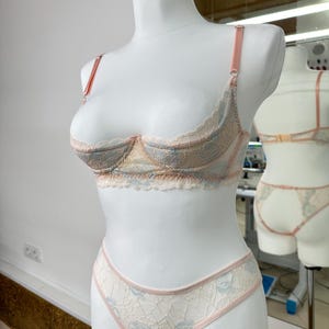 May include: A two-piece lingerie set featuring a lace bra and matching panties. The set is crafted from cream-colored lace with light blue floral patterns and accented with thin, pale pink straps and trim along the edges.