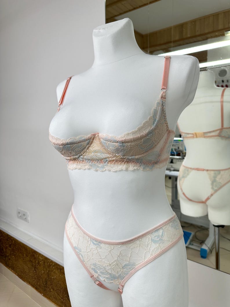 May include: A two-piece lingerie set featuring a lace bra and matching panties. The set is crafted from cream-colored lace with light blue floral patterns and accented with thin, pale pink straps and trim along the edges.