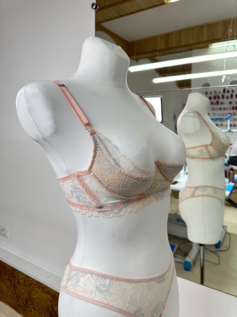 May include: A beige and light blue lace lingerie set displayed on a mannequin. The bra and matching bottoms feature delicate floral lace detailing with pink trim. The set is designed with adjustable straps and a classic silhouette.