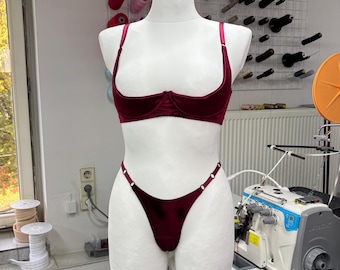 May include: A burgundy lingerie set displayed on a mannequin. The set includes a demi-cup bra with adjustable straps and a matching thong. The fabric appears to be a soft velvet. The bra and thong are a deep red color.