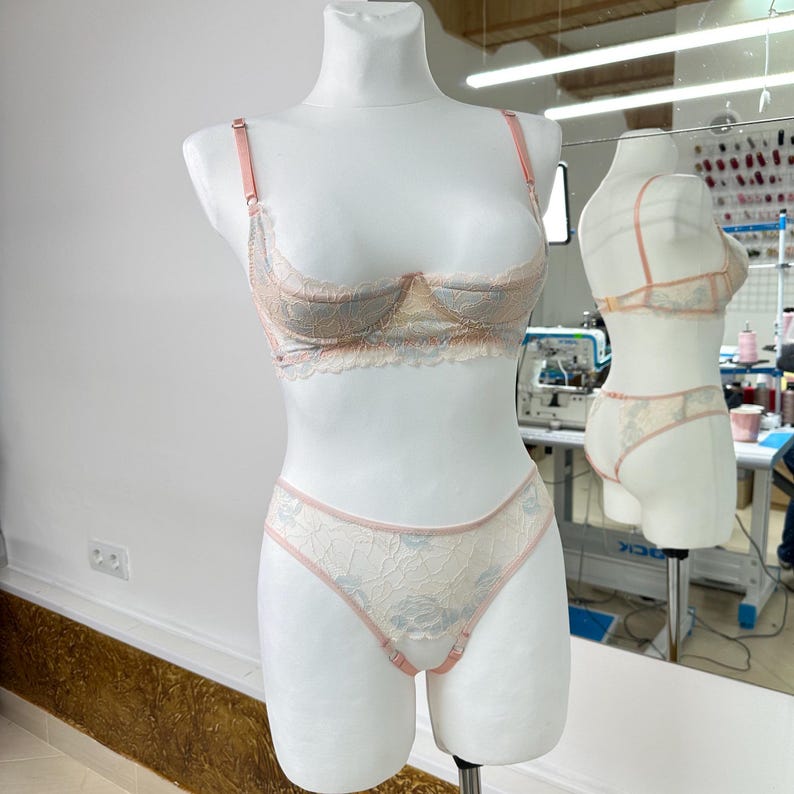 May include: A delicate lingerie set displayed on a mannequin. The set includes a balconette bra and matching briefs, both in a soft, floral lace pattern with pink and blue accents. The bra has adjustable straps. The overall color palette is pastel.