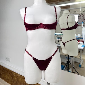 Op de afbeelding: Een bordeauxrode fluwelen lingerieset op een mannequin. De set bestaat uit een demi-cup bh met verstelbare bandjes en een bijpassende string. De string heeft decoratieve metalen ringen. De bh en string zijn diep bordeaux van kleur.