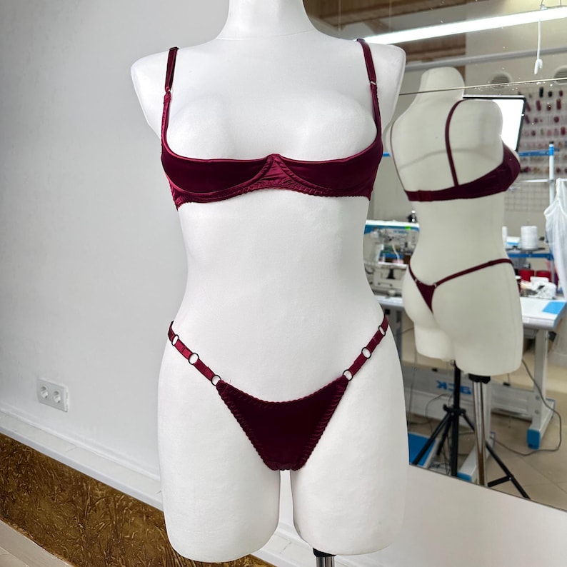 Peut inclure: Ensemble de lingerie en velours bordeaux pr&eacute;sent&eacute; sur un mannequin. L'ensemble comprend un soutien-gorge demi-bonnet avec des bretelles r&eacute;glables et un string assorti. Le string comporte des anneaux m&eacute;talliques d&eacute;coratifs. Le soutien-gorge et le string sont de couleur bordeaux fonc&eacute;.