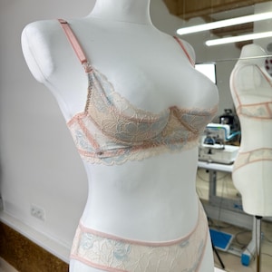 May include: A two-piece lingerie set featuring a lace underwire bra and matching panties. The set is crafted from cream-colored lace with light blue floral patterns and accented with soft pink trim, adjustable straps, and metal hardware.
