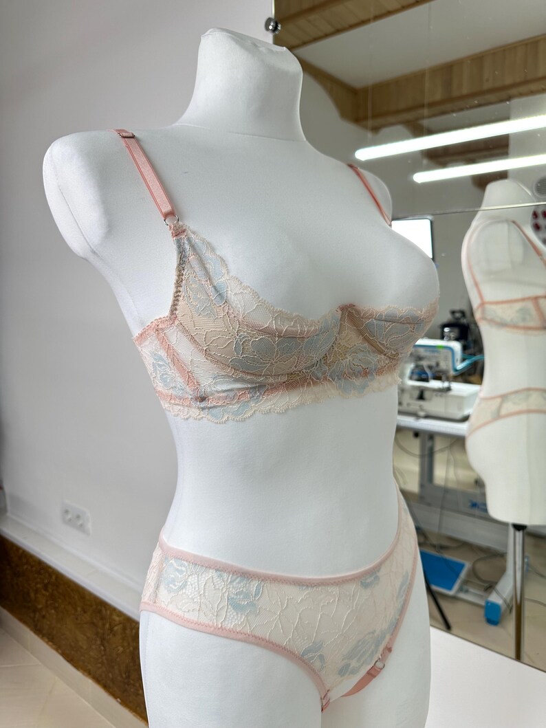 May include: A two-piece lingerie set featuring a lace underwire bra and matching panties. The set is crafted from cream-colored lace with light blue floral patterns and accented with soft pink trim, adjustable straps, and metal hardware.