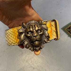 Puede incluir: Un cinturón con una correa texturizada de patrón de piel de serpiente en amarillo y blanco, y una hebilla metálica grande en tono bronce antiguo con forma de cabeza de león detallada, con una melena prominente y rasgos faciales.