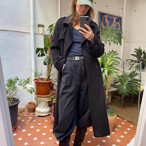 May include: A person wearing a black trench coat, navy blue pants, and brown suede boots. They are also wearing a green baseball cap with a white logo. The person is holding a phone and taking a picture in front of a mirror.