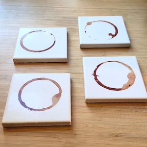 May include: Four square white ceramic coasters with brown coffee ring stains. The coasters are arranged on a light brown wooden surface. Each coaster has a unique coffee stain pattern.