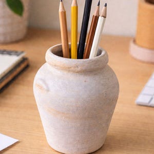 Może przedstawiać: Beżowy ceramiczny pojemnik na ołówki o teksturowanej powierzchni, zawierający kilka zaostrzonych ołówków w różnych kolorach: żółtym, czarnym, brązowym i białym. Ołówki znajdują się w małym wazonie w stylu rustykalnym, ustawionym na drewnianym biurku.