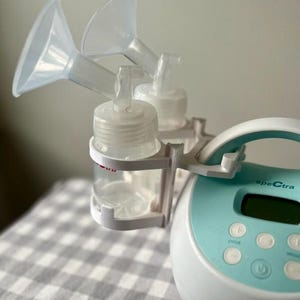 May include: A white and blue electric breast pump with a digital display and buttons. The pump has two clear bottles with white caps and two clear silicone funnels attached.