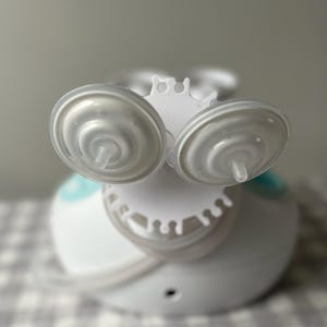 May include: White electric breast pump with two clear silicone breast shields and a white base. The pump has a blue accent and a white drip catcher.