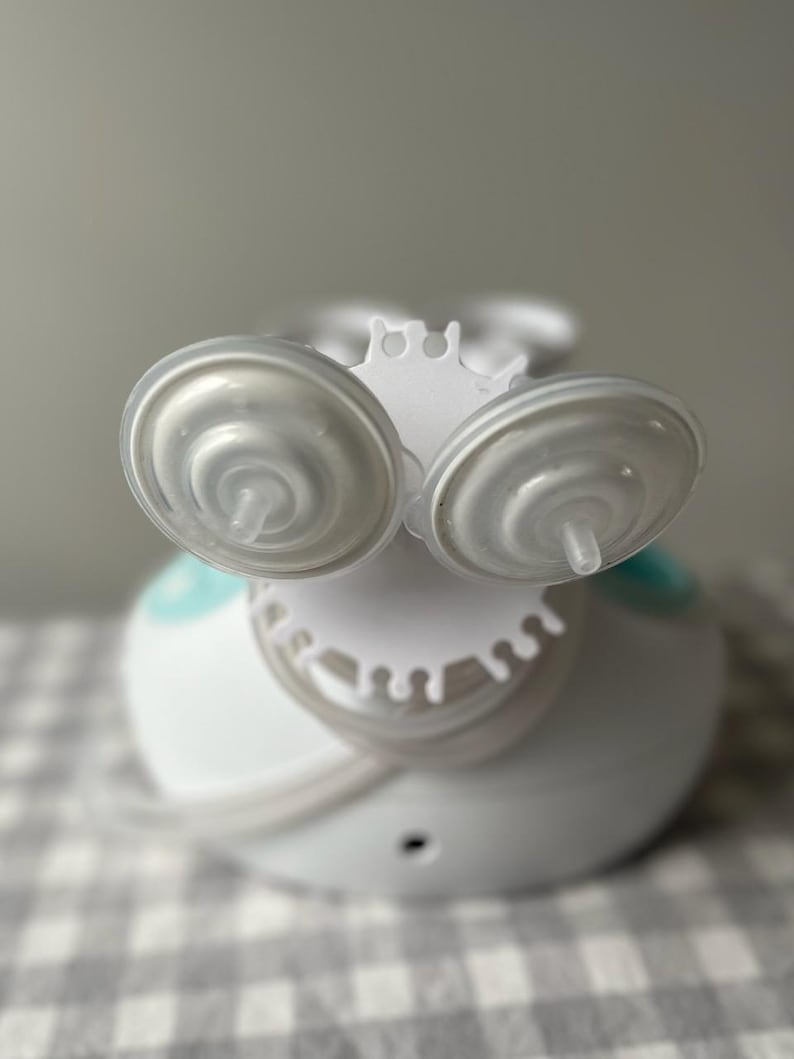 May include: White electric breast pump with two clear silicone breast shields and a white base. The pump has a blue accent and a white drip catcher.