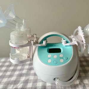 May include: A white electric breast pump with a blue control panel and two clear bottles. The pump has a handle and two clear tubes connected to the bottles. The pump is on a white and gray checkered surface.