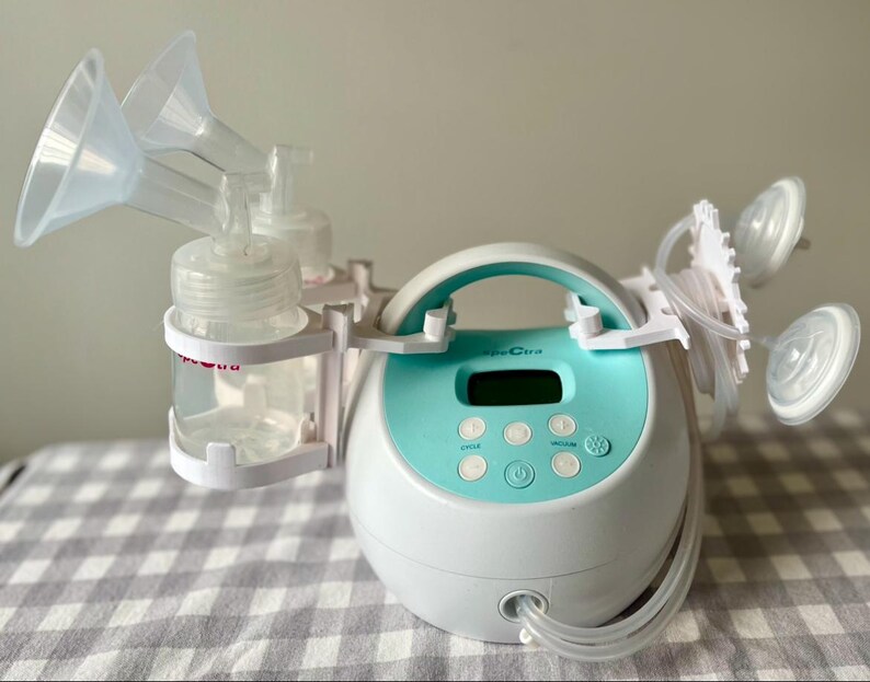May include: A white electric breast pump with a blue control panel and two clear bottles. The pump has a handle and two clear tubes connected to the bottles. The pump is on a white and grey checkered surface.
