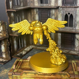 Op de afbeelding: Een goudkleurig geschilderd miniatuurfiguur van een gevleugelde humanoïde krijger in vlucht, zwevend boven een ronde basis door een gebeeldhouwde pluim van rook of uitlaatgassen. Het figuur heeft een gespierd pantser en grote, gespreide vleugels.
