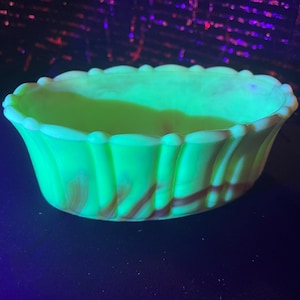 Op de afbeelding: Een groene glazen kom met een geribbelde rand en een swirl-patroon. De kom is gemaakt van uraniumglas en gloeit onder een zwart licht.