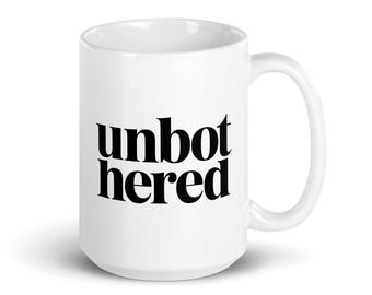 Könnte beinhalten: Weißer Keramikbecher mit dem Wort "unbothered" in schwarzer, fetter, serifenloser Schrift. Der Becher hat einen gebogenen Henkel und eine zylindrische Form.