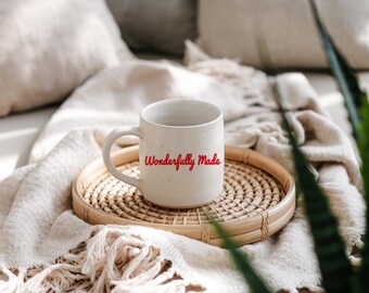 Könnte beinhalten: Weiße Keramiktasse mit einer roten Aufschrift "Wonderfully Made" auf einem geflochtenen Weidentablett. Die Tasse steht auf einer weißen Decke mit Fransen.