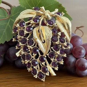 May include: A gold-toned brooch in the shape of a grape cluster, adorned with purple rhinestones. The brooch features detailed leaves and stems. The image includes real grapes and leaves, creating a naturalistic display.