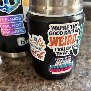May include: Black insulated water bottle with various stickers. Stickers include phrases like "You're the good kind of weird, I value that" and "Surviving Deadlines". The bottle has a stainless steel top and is sitting on a brown surface.