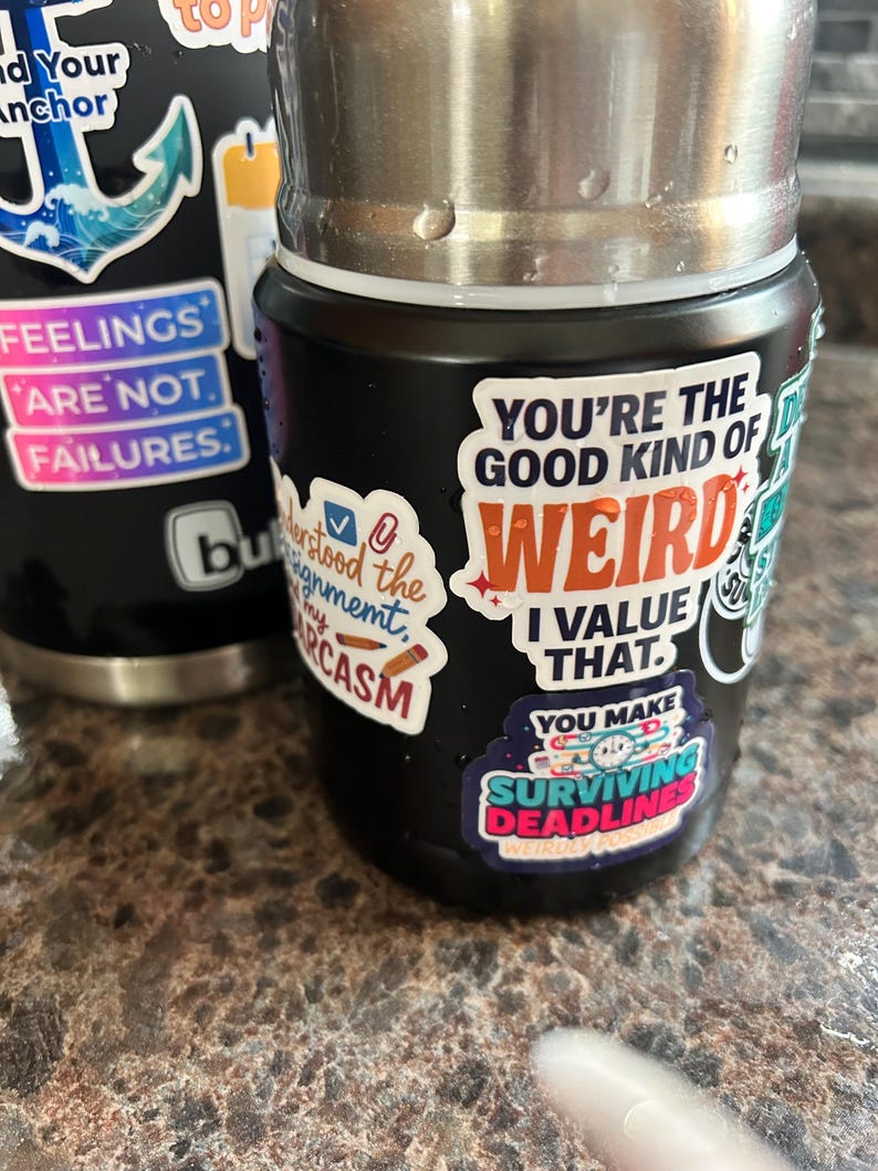 May include: Black insulated water bottle with various stickers. Stickers include phrases like "You're the good kind of weird, I value that" and "Surviving Deadlines". The bottle has a stainless steel top and is sitting on a brown surface.