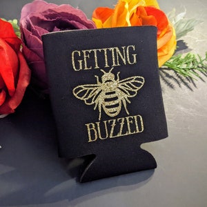 May include: Black drink koozie with gold glitter text and a bee graphic. The text reads "GETTING BUZZED". The koozie is surrounded by red, purple, and yellow flowers.